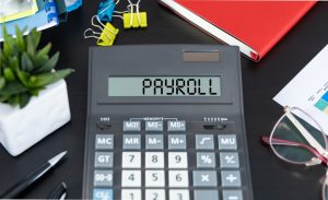 On the dark table there is a black calculator with the text PAYROLL on the scoreboard, a pen and other office tools. Business concept